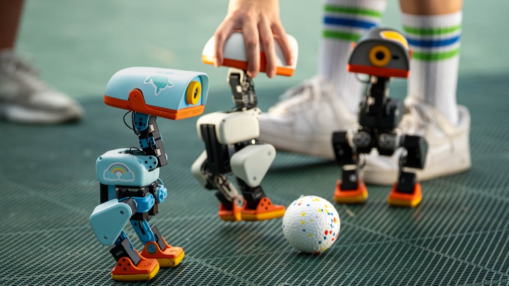 Three MicroDuck robots beside a football, with a hand and shoes showing their scale